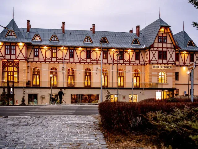 A large, multi-story building with timber framing, lit windows, and a sign reading "Hotel Lomnice" is seen at dusk with a cobblestone walkway and shrubs in the foreground.