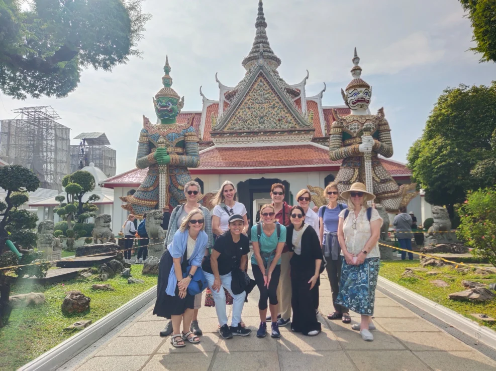 A group of ten people pose for a photo in front of ornate guardian statues at a temple with traditional architecture and landscaped gardens.