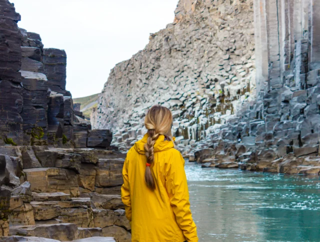 A person in a yellow jacket stands by a river surrounded by basalt rock columns and cliffs.