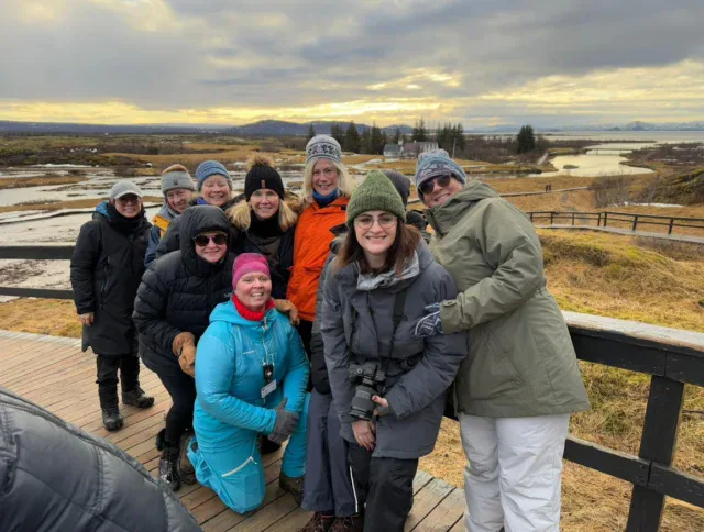A group of people dressed in winter outdoor clothing pose and smile on a wooden walkway in a scenic, open landscape under a cloudy sky.
