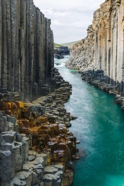 A person stands on rocky basalt columns beside a turquoise river flowing between tall, vertical cliffs under a partly cloudy sky.