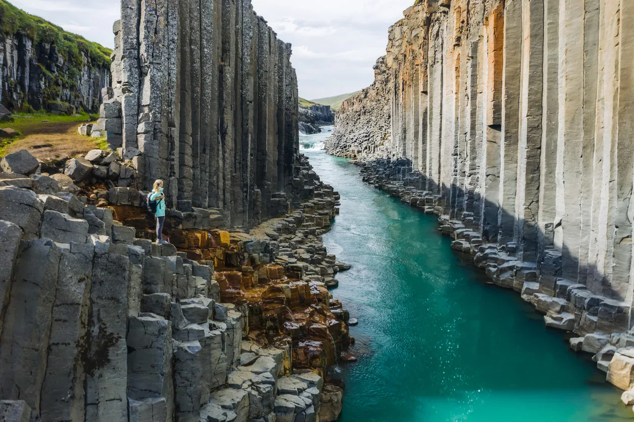 A person stands on rocky basalt columns beside a turquoise river flowing between tall, vertical cliffs under a partly cloudy sky.