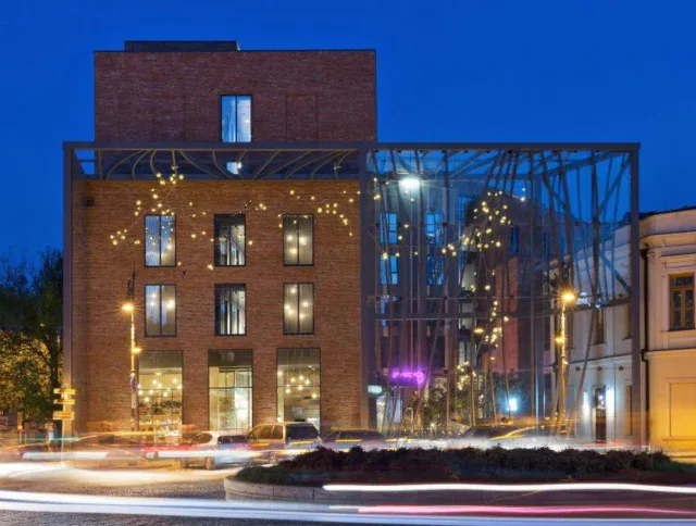 A modern brick building with large windows and a steel-framed glass extension illuminated at night, with light trails from passing cars in the foreground.