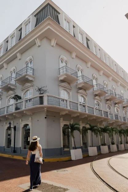 A woman in a hat walks along a brick street past a large, white colonial-style building with arched windows and balconies on a sunny day.