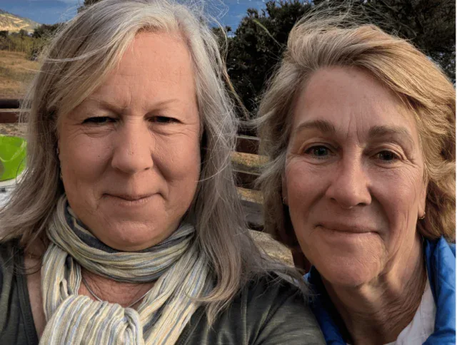 Two older women with gray hair taking a selfie outdoors, with trees and mountains visible in the background.