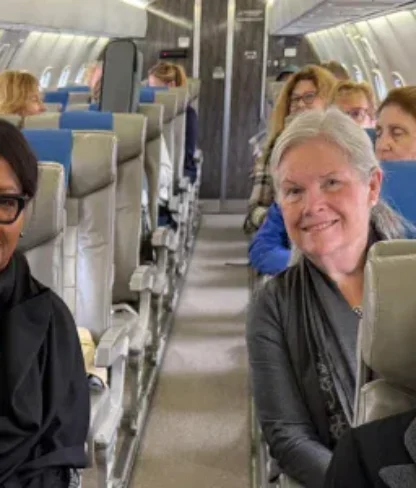 Passengers, mostly older women, sit in pairs on a small commercial airplane with blue and gray seats, facing forward and smiling at the camera.