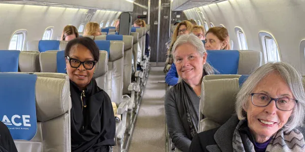 Passengers, mostly older women, sit in pairs on a small commercial airplane with blue and gray seats, facing forward and smiling at the camera.