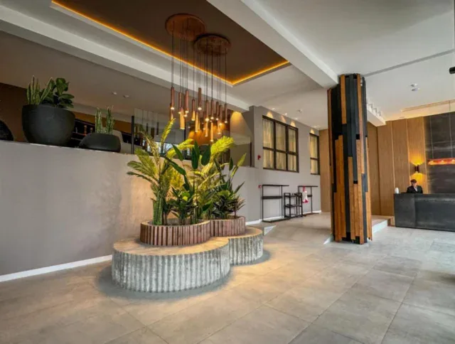 Modern hotel lobby with potted plants on a circular bench, pendant lights above, tiled floors, and a reception desk in the background.