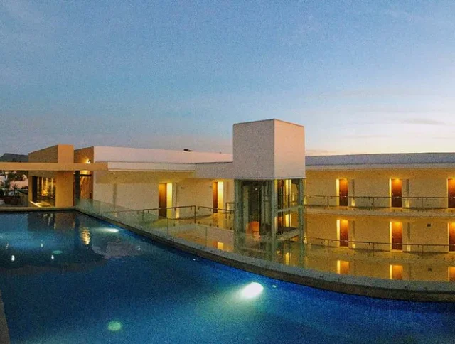 Modern building with illuminated balconies and a rooftop swimming pool at dusk, reflecting the structure and evening sky.