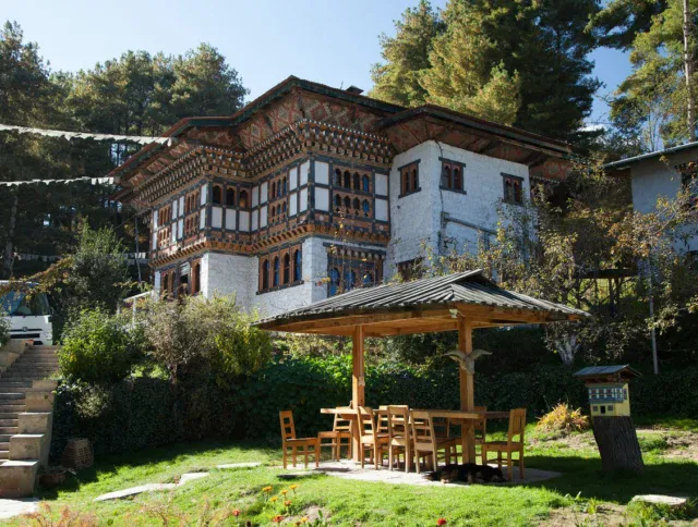 A traditional Bhutanese building with ornate woodwork stands behind a wooden outdoor seating area, surrounded by greenery and trees.