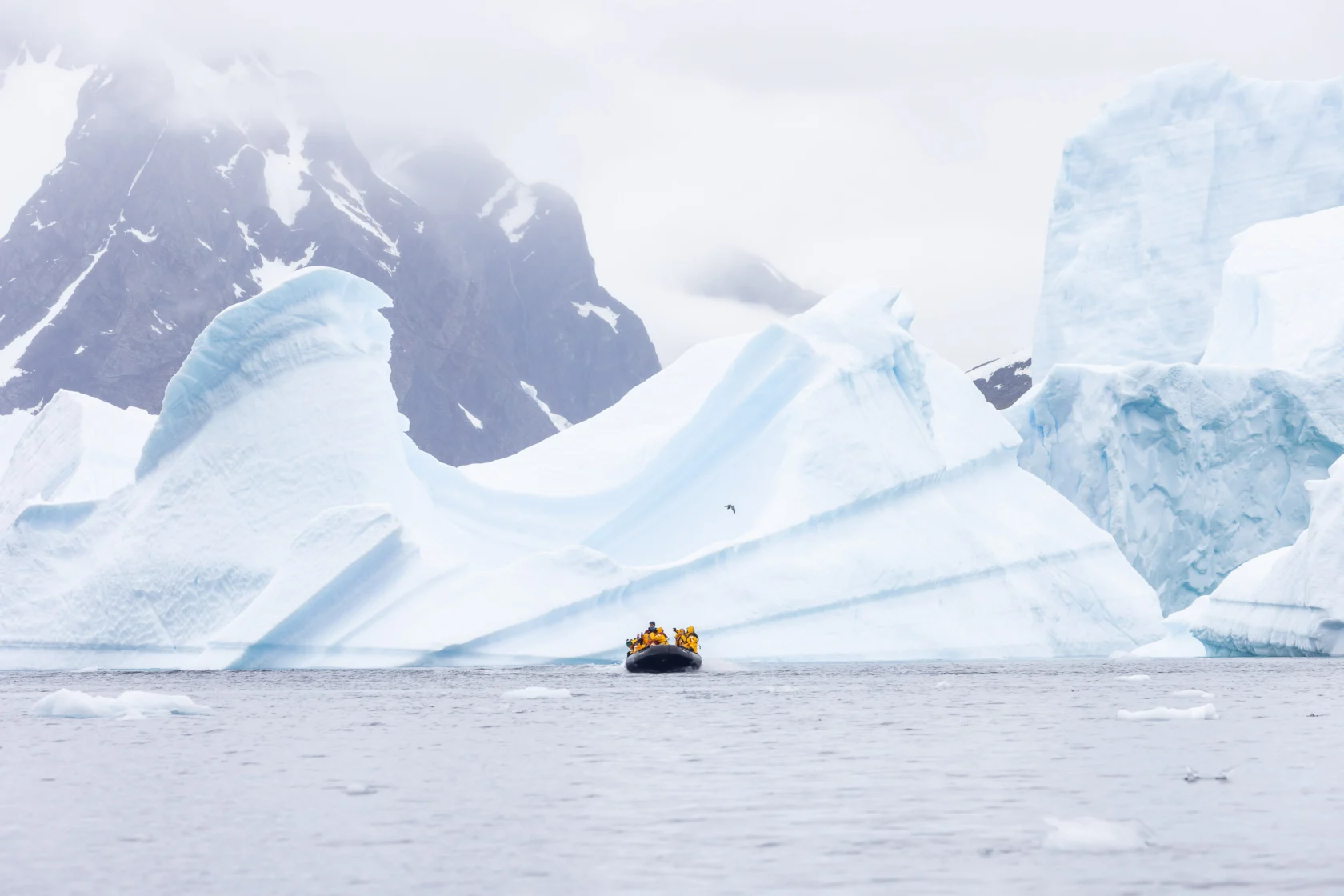A small inflatable boat with people in yellow jackets travels near large icebergs on calm, gray water with snow-covered mountains in the background.
