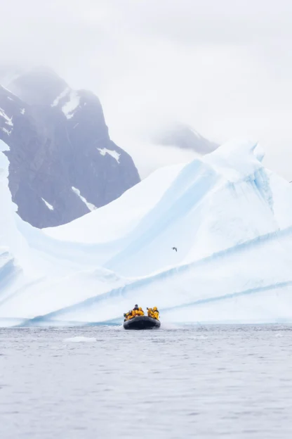 A small inflatable boat with people in yellow jackets travels near large icebergs on calm, gray water with snow-covered mountains in the background.