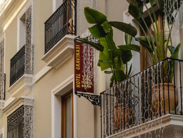 A hotel building facade with a "Hotel Gravina 51" sign, balconies decorated with potted green plants, and hanging string lights.