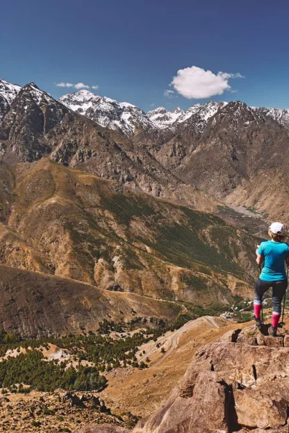 A person stands on a rocky outcrop overlooking a vast mountain landscape with snow-capped peaks under a clear blue sky.
