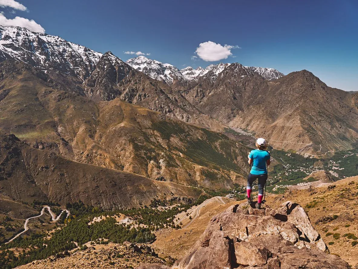 A person stands on a rocky outcrop overlooking a vast mountain landscape with snow-capped peaks under a clear blue sky.