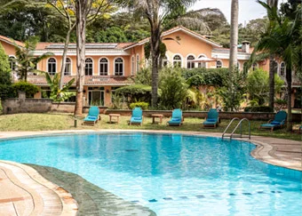 A curved outdoor swimming pool with blue lounge chairs around it, set in front of a large two-story house surrounded by trees and greenery.