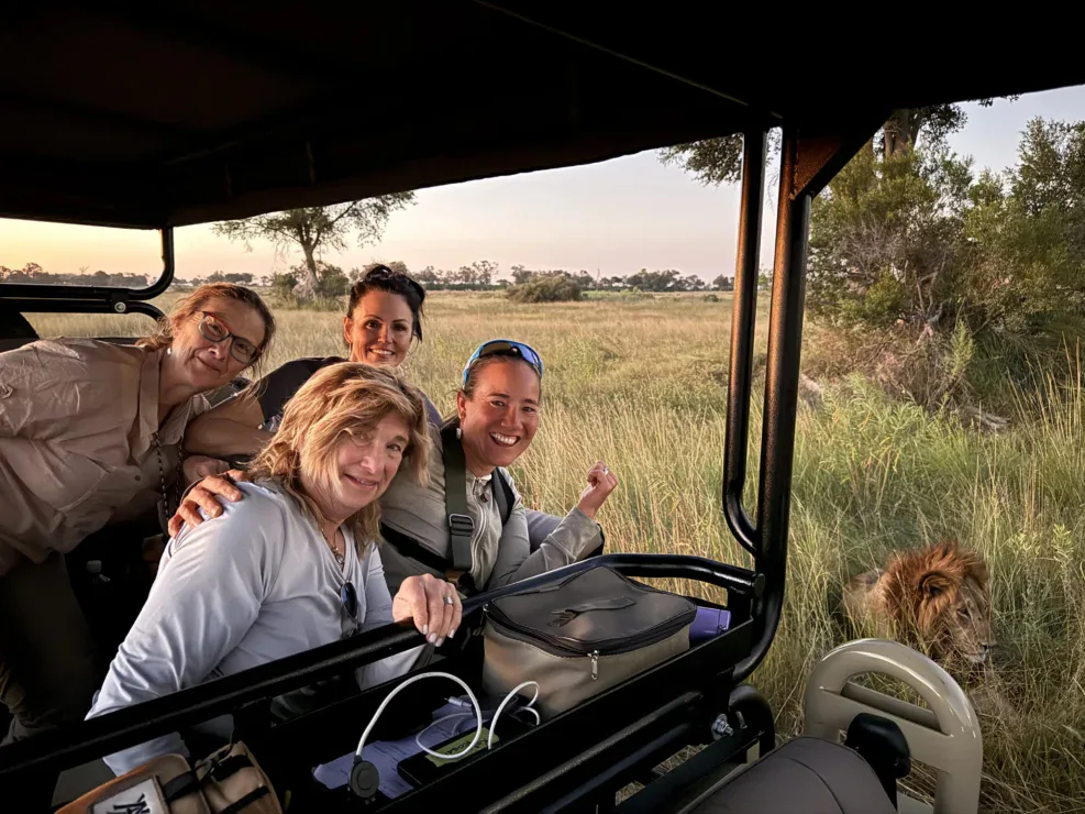 Four people in a safari vehicle smiling and posing for a photo with a male lion lying in the grass nearby.