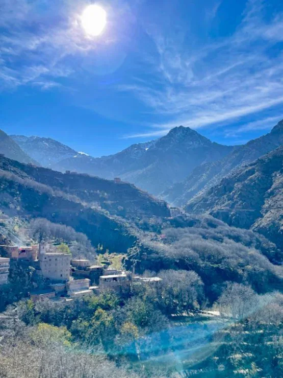 Sun shines over a mountainous landscape with scattered buildings, leafless trees, and a clear blue sky with wispy clouds.