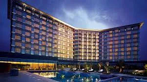 A modern multi-story hotel building with illuminated windows, viewed at dusk, featuring a lit outdoor swimming pool in the foreground.