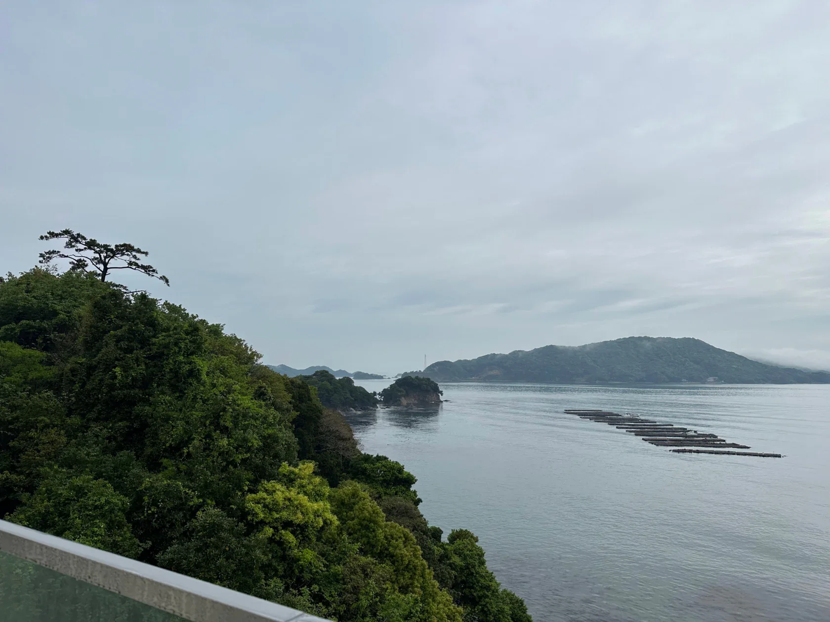 View of a calm coastal landscape with green forested cliffs, small islands, floating platforms on the water, and a cloudy sky.