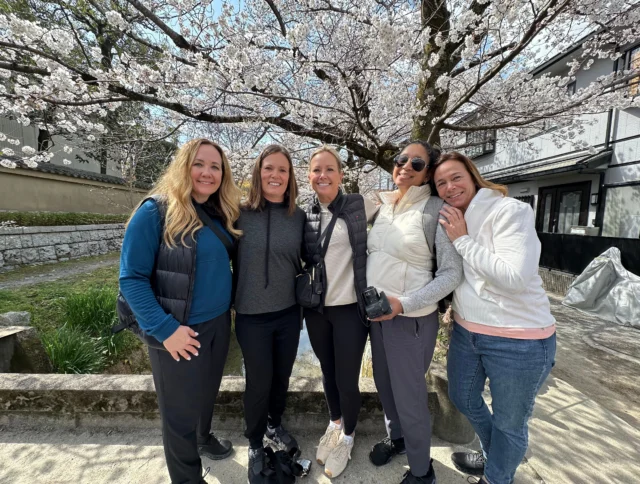 Five women stand together smiling under blooming cherry blossom trees on a sunny day, with buildings and greenery in the background.
