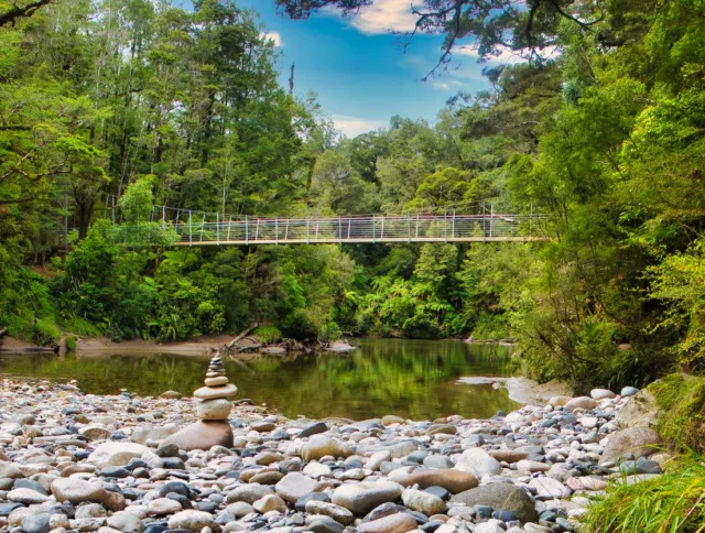 Suspension bridge crosses a calm river surrounded by dense green forest, with stacked stones and smooth rocks in the foreground.