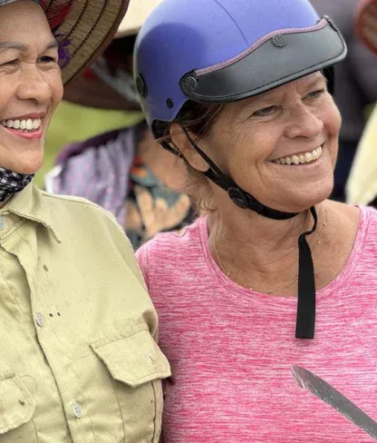 Two women smiling, one in a conical hat flashing a peace sign, the other in a helmet and pink shirt holding a tool, with other people in the background.