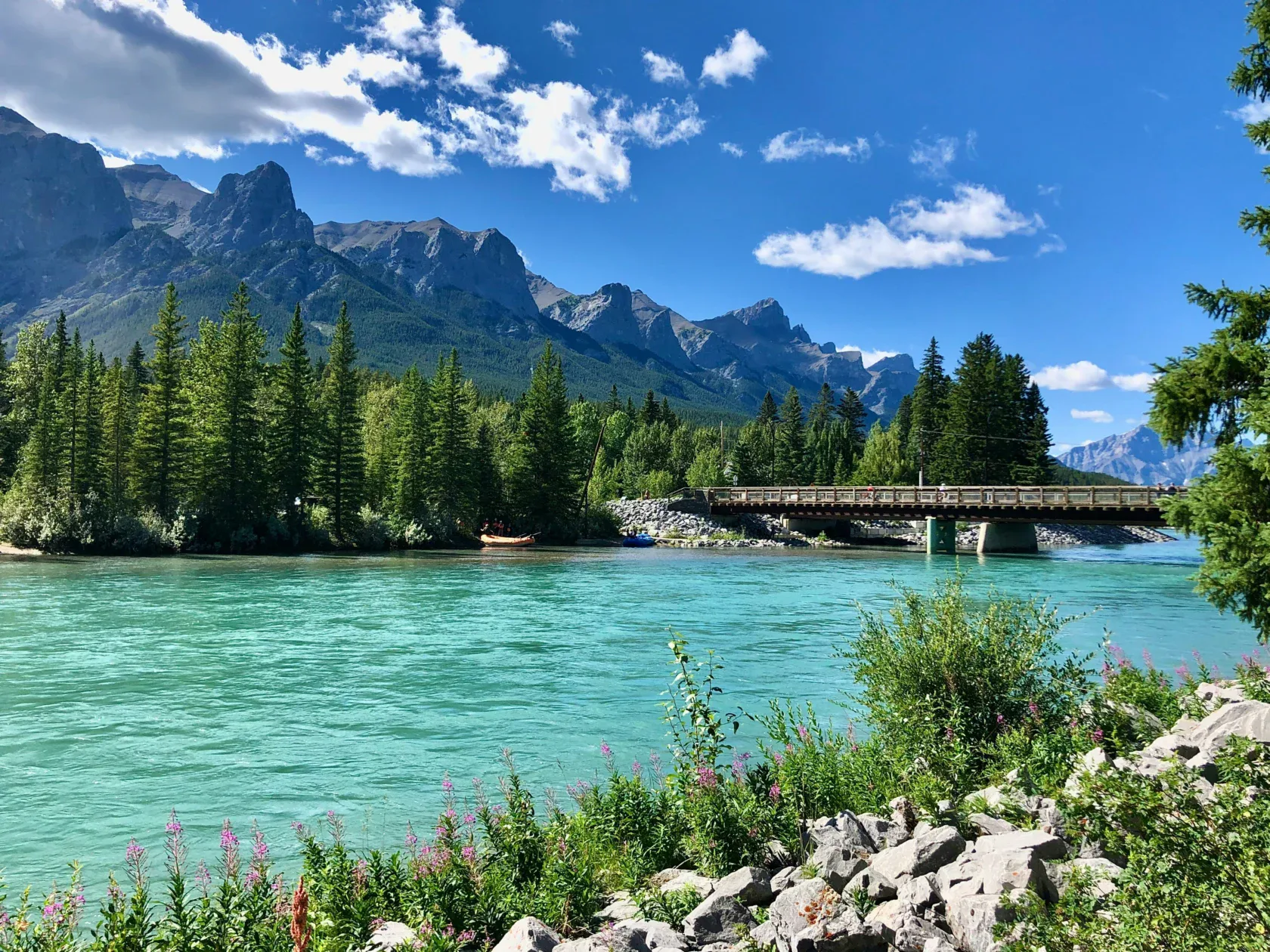 A turquoise river flows beneath a wooden bridge, surrounded by evergreen trees and rocky banks, with mountains and a blue sky in the background.