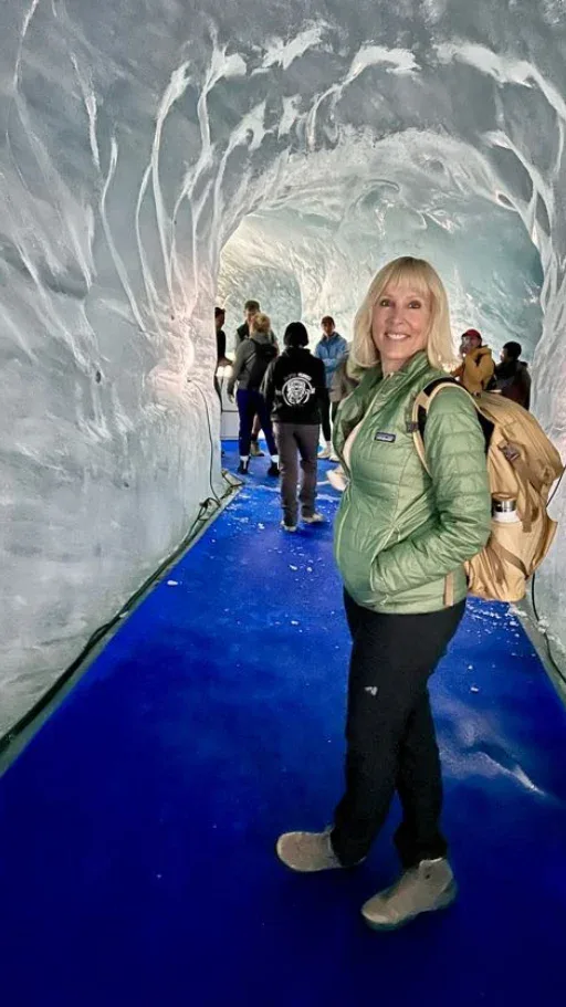 A woman with a backpack stands in a tunnel made of ice with a blue walkway and several people in the background.