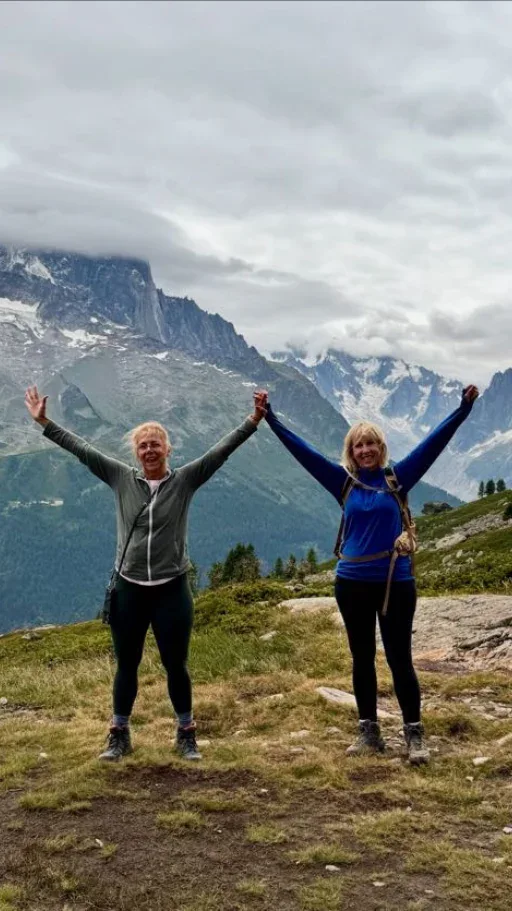 Two women stand on a grassy mountain trail with arms raised, holding hands, with snow-capped mountains and cloudy sky in the background.