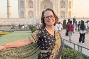 A woman wearing a traditional Indian saree stands smiling in front of the Taj Mahal, with tourists and gardens visible in the background.