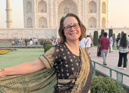 A woman wearing a traditional Indian saree stands smiling in front of the Taj Mahal, with tourists and gardens visible in the background.