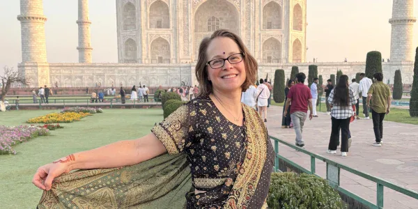 A woman wearing a traditional Indian saree stands smiling in front of the Taj Mahal, with tourists and gardens visible in the background.