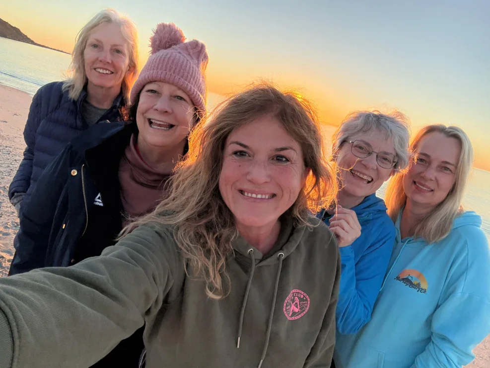 Five women stand together on a beach at sunset, smiling for a group selfie. The ocean and a headland are visible in the background.