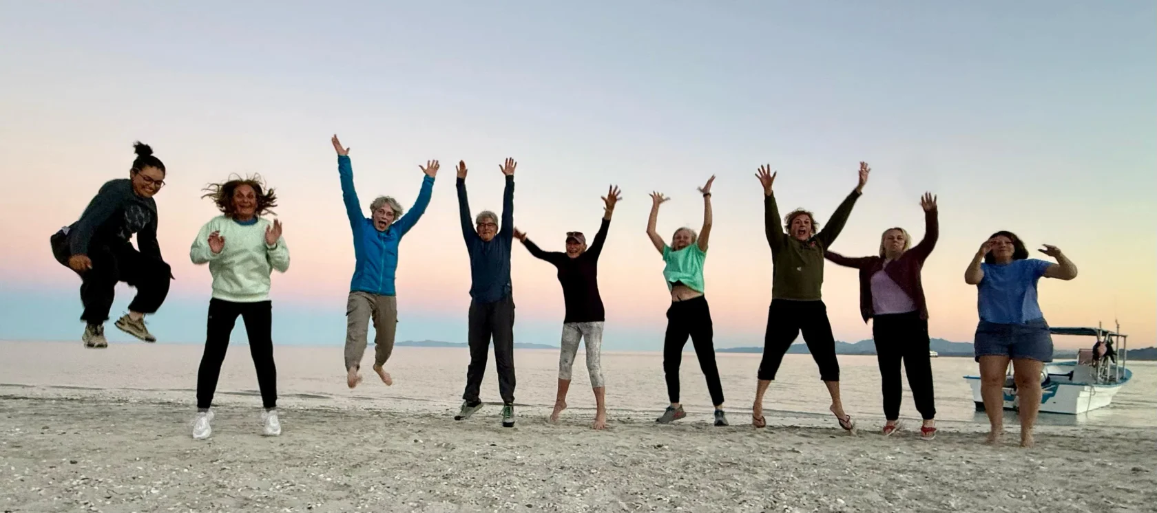 A group of nine people jump in the air on a sandy beach at sunset, with the sea and a boat in the background.