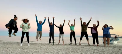 A group of nine people jump in the air on a sandy beach at sunset, with the sea and a boat in the background.