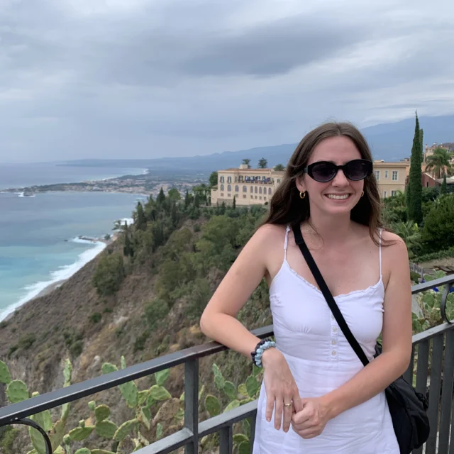 Woman in white dress and sunglasses standing by a railing overlooking a coastal town with the sea and hills in the background under a cloudy sky.