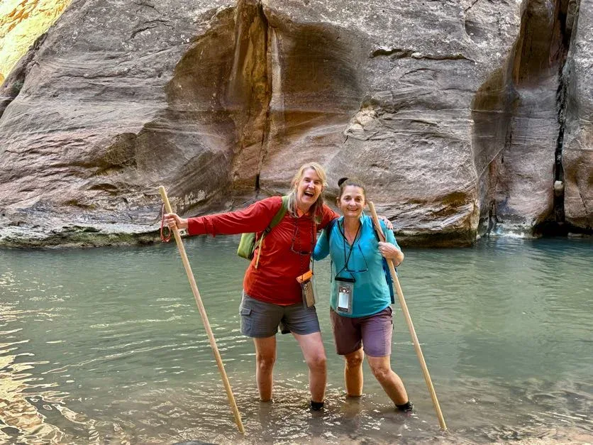 Two people stand in shallow river water with walking sticks, smiling at the camera. They are outdoors, with a large rock wall in the background.