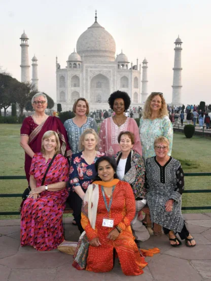 A group of ten women pose for a photo in front of the Taj Mahal, with some seated and others standing, during daylight.