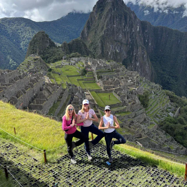 Three women pose in a tree yoga position on a grassy hill overlooking the ancient ruins of Machu Picchu with mountains in the background.
