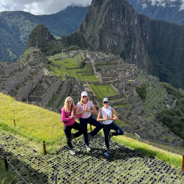 Three women pose in a tree yoga position on a grassy hill overlooking the ancient ruins of Machu Picchu with mountains in the background.