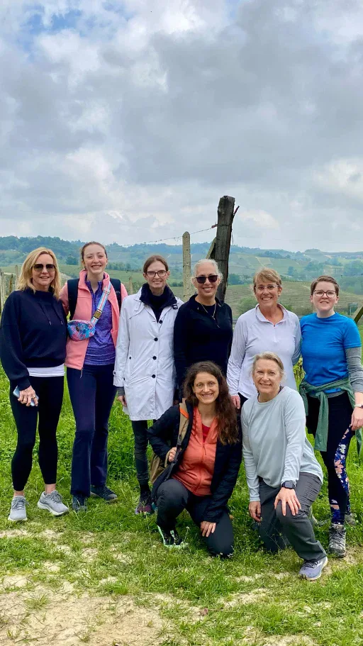 Eight women pose together outdoors on a grassy area with hills and a cloudy sky in the background.