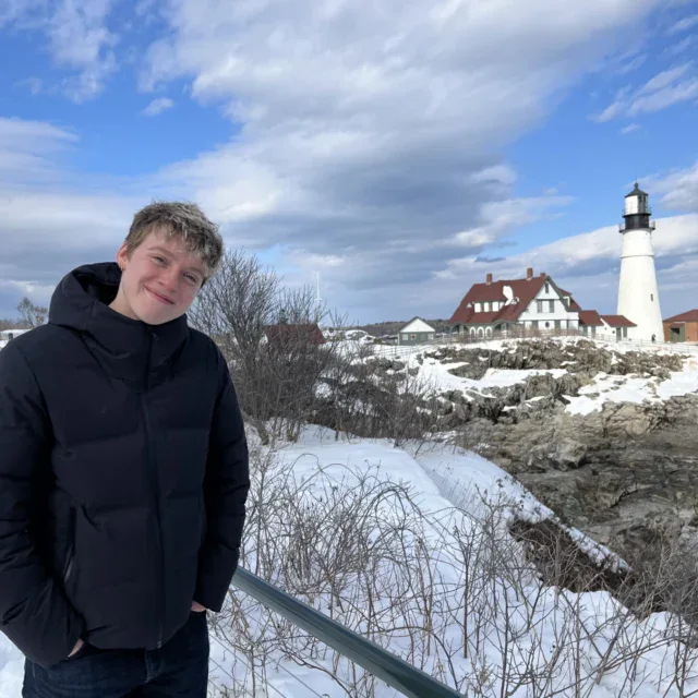 Person in a black jacket stands outdoors in front of a snow-covered landscape with a lighthouse and buildings in the background under a cloudy sky.