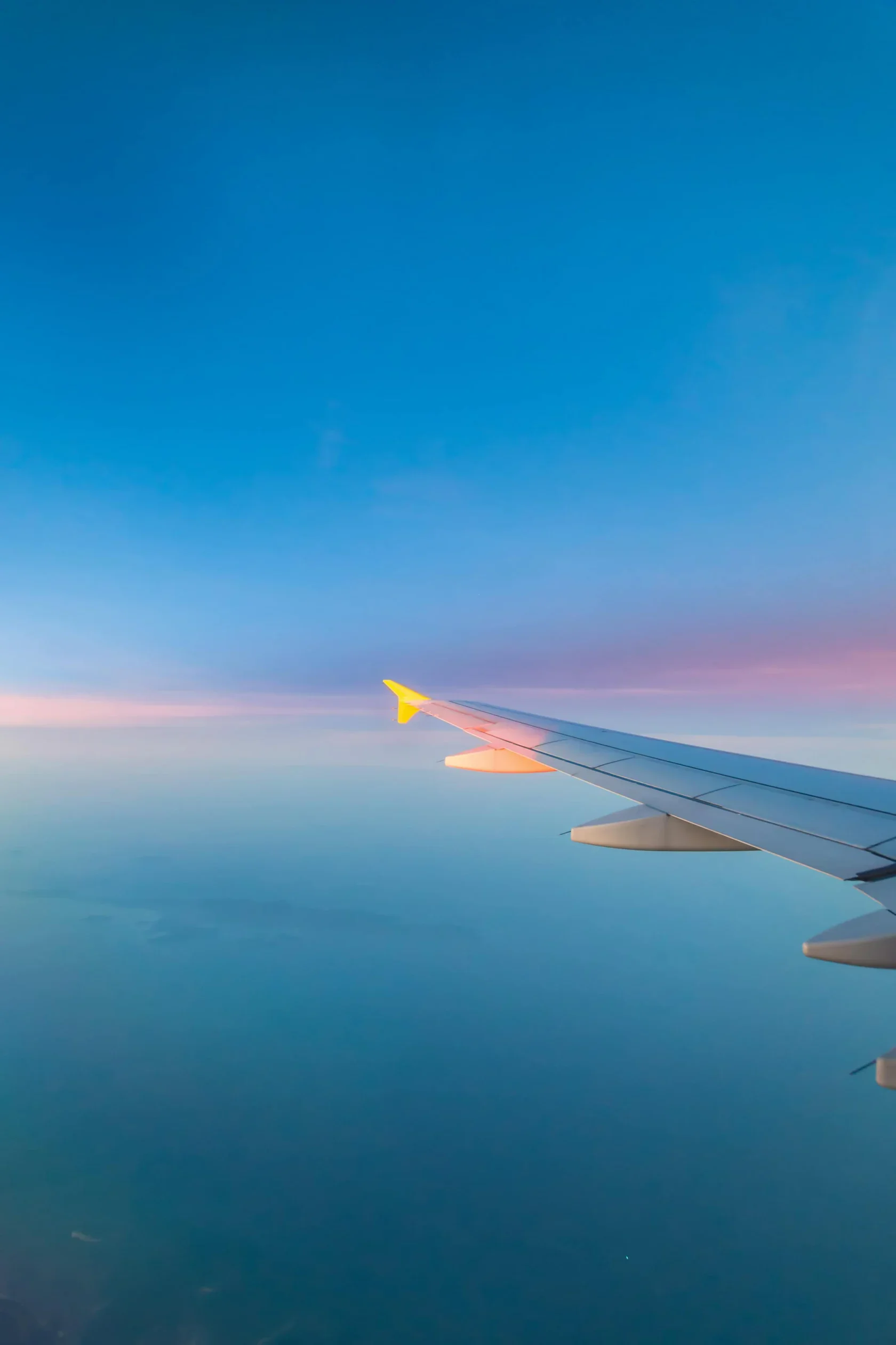 View from an airplane window showing the wing against a blue sky with pink and purple clouds at sunrise or sunset.