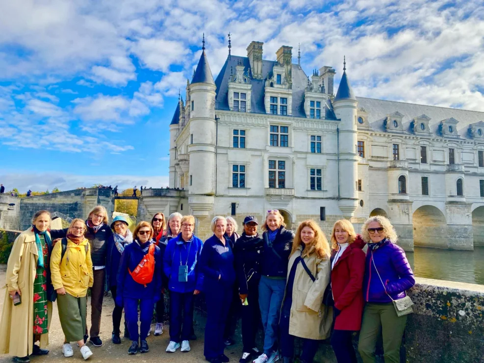 A group of people pose for a photo in front of a large, historic chateau with white stone walls and multiple turrets under a blue sky.