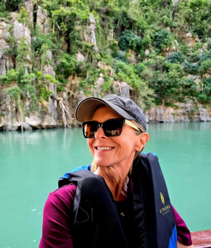 Woman wearing a cap, sunglasses, and life vest smiles while sitting on a boat; turquoise water and rocky, forested cliffs in the background.