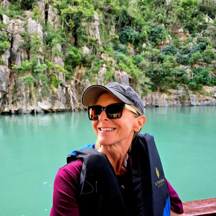 Woman wearing a cap, sunglasses, and life vest smiles while sitting on a boat; turquoise water and rocky, forested cliffs in the background.