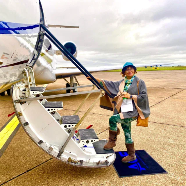 A person wearing a blue hat and holding a boarding pass stands on the steps of a private jet on an airport tarmac under a cloudy sky.