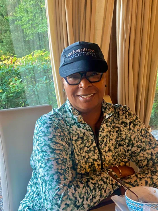 A woman wearing glasses and a black "Adventure Women" cap sits at a table near a window with greenery outside, smiling with her arms crossed.