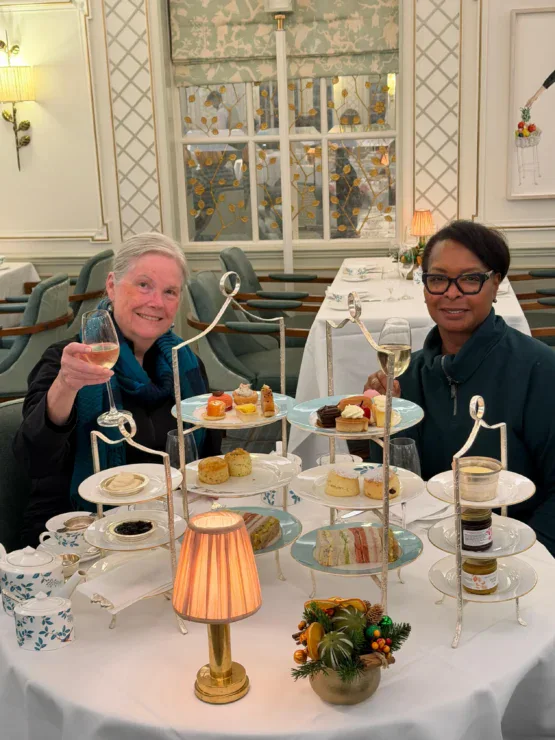 Two women sit at a table set for afternoon tea with tiered trays of pastries, sandwiches, and desserts; one woman holds up a glass and smiles at the camera.